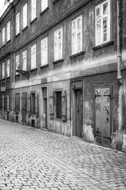 Gri tonlu bir görüntü Avrupa 'daki bir sokak manzarasını yakalıyor. Binanın alt katında kapılar ve pencereler var, bazıları graffitiyle kaplı. Yukarıda, pencereler üst katlarda. Sokaklar kaldırım taşlarıyla kaplı..