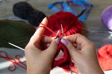 Kadın elleri şiş ve kırmızı iplikle çorap örüyor. Kırmızı iplikler, leylak ve koyu yeşil renk. Gri ahşap arka plan. Ellerini yaklaştır.