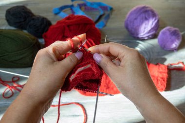 Kadın elleri şiş ve kırmızı iplikle çorap örüyor. Kırmızı iplikler, leylak ve koyu yeşil renk. Gri ahşap arka plan. Ellerini yaklaştır.