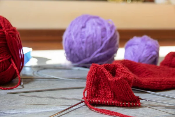 Sock in the process of knitting with needles and yarn of red color on a gray wooden table. Clews threads of red and lilac color on a blurred background. Close-up