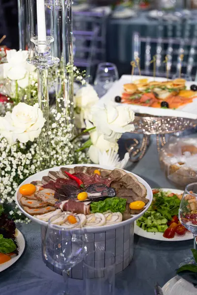 Assortment of cured meats garnished with chili peppers and parsley, artfully arranged on a platter. Placed on a table decorated with flowers and candles, creating an inviting centerpiece.