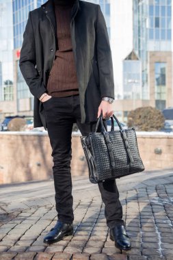 Siyah ceketli balıkçı yaka kazaklı, deri çantalı, ofisin yanında duran bir iş adamı. Kot ayakkabı giyiyor. Düşük açı görünümü, bulanık arkaplan.