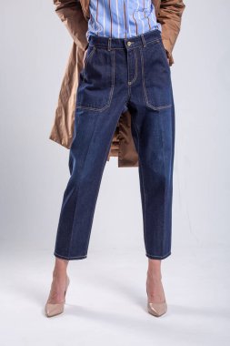 Yüksek bel kotlu, mavi gömlekli, kahverengi trençkotlu ve beyaz arka planda çıplak topuklu genç bir kadın..