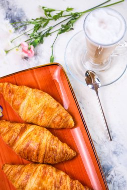 Üzerinde kruvasan ve mermer masa manzaralı kahve olan enfes bir kahvaltı. Kruvasanlar turuncu bir tabakta altın kahverengidir, köpüklü kahve..