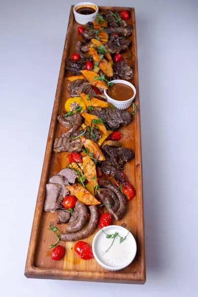 A long wooden platter filled with an assortment of meats and vegetables, including steak, sausage, chicken, potatoes, carrots, and peppers.