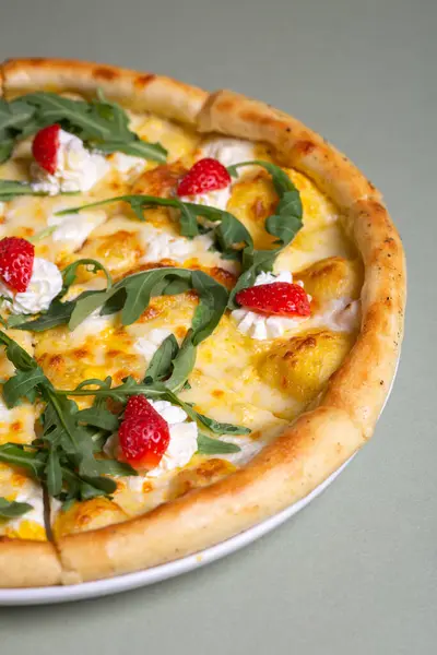 A delicious pizza with strawberries, cream cheese, and arugula on a white plate. The pizza is isolated on a green background. The pizza is perfect for a summer party or a picnic.