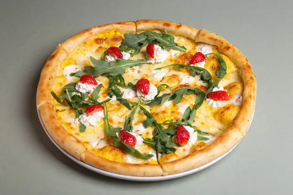 A delicious pizza with strawberries, cream cheese, and arugula on a white plate. The pizza is isolated on a green background. The pizza is perfect for a summer party or a picnic.