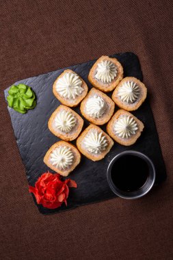 Deep-fried sushi roll with cream cheese and salmon, served on a black plate with soy sauce and wasabi paste. Topped with creamy sauce. clipart