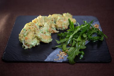 Yakından çekilmiş, bol yağda kızartılmış avokado karidesi, siyah taş tabakta roka, deniz ürünleri severler için mükemmel..