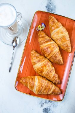 Üzerinde kruvasan ve mermer masa manzaralı kahve olan enfes bir kahvaltı. Kruvasanlar turuncu bir tabakta altın kahverengidir, köpüklü kahve..