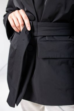 Bir kadın siyah bir ceketin kemerini ince ve klasik yakalı olarak ayarlıyor. Şık ve sofistike bir iş kıyafeti yaratıyor..