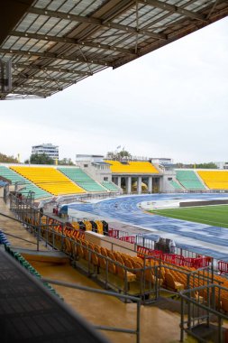 Mavi raylı parlak sarı stadyum koltukları. Tamire ihtiyacı olan eski stadyum. Çatlak beton ve soluk boya görünür..