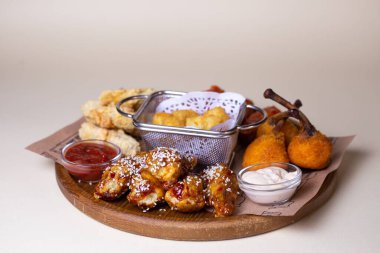 A variety of fried chicken wings, potato balls, and cheese sticks with dipping sauces on a wooden platter. clipart
