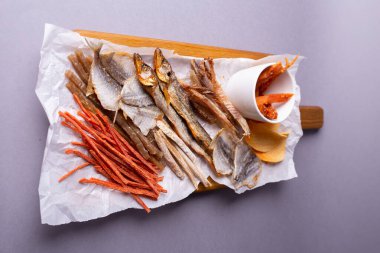 Tahta masadaki beyaz kağıt üzerinde çeşitli kurutulmuş balıklar, balık derisi ve meyve atıştırmalıkları, canlı bir mutfak görüntüsü yaratıyor..