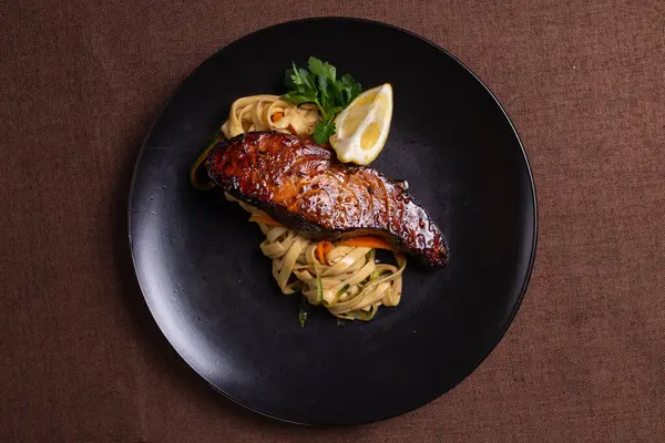 stock image A mouthwatering and nutritious dish featuring grilled salmon, lemon, and fresh vegetables on a sleek black plate.