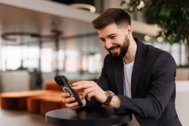 Başarılı yatırımcı ofis içinde çalışır, iş adamı takım elbiseli iş adamı pencere kenarındaki telefonu kullanır, adam gülümser ve akıllı telefondan iyi haberler okur..