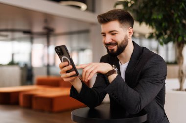 Başarılı yatırımcı ofis içinde çalışır, iş adamı takım elbiseli iş adamı pencere kenarındaki telefonu kullanır, adam gülümser ve akıllı telefondan iyi haberler okur..