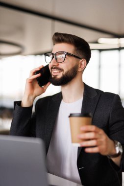 Gülümseyen genç işadamı iş yerinde kahve molası veriyor ve modern beyaz ofiste dizüstü bilgisayarla konuşurken cep telefonuyla konuşuyor.