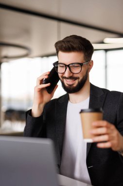 Gülümseyen genç işadamı iş yerinde kahve molası veriyor ve modern beyaz ofiste dizüstü bilgisayarla konuşurken cep telefonuyla konuşuyor.