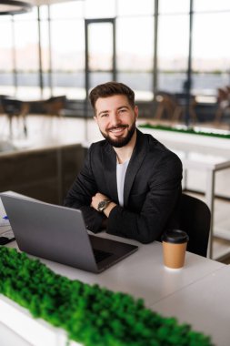 Şık, kısa sakallı çekici bir işletmeci. Ofis masasında oturmuş, penceresinden iyimser bir ifadeyle dışarı bakıyor.