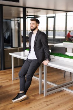 Modern ofiste duran iş adamının portresi. Başarılı bir yetişkin girişimci resmi giysiler içinde kameraya zevkle bakıyor. Gözlüklü ve sakallı, kendine güvenen bir adam..