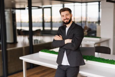 Modern ofiste duran iş adamının portresi. Başarılı bir yetişkin girişimci resmi giysiler içinde kameraya zevkle bakıyor. Gözlüklü ve sakallı, kendine güvenen bir adam..