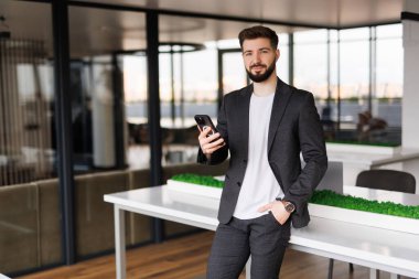 Genç, gülümseyen bir adam elinde bir telefonla, bir ofis binasının önünde yürüyor, mutlu bir şekilde akıllı telefondan bir uygulama kullanıyor, mesajları, tipleri okuyor ve internette geziniyor..