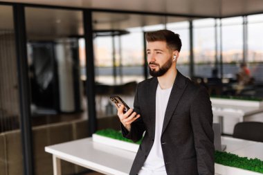 Genç, gülümseyen bir adam elinde bir telefonla, bir ofis binasının önünde yürüyor, mutlu bir şekilde akıllı telefondan bir uygulama kullanıyor, mesajları, tipleri okuyor ve internette geziniyor..