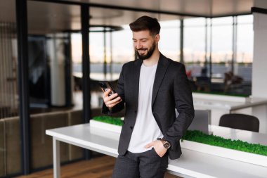 Genç, gülümseyen bir adam elinde bir telefonla, bir ofis binasının önünde yürüyor, mutlu bir şekilde akıllı telefondan bir uygulama kullanıyor, mesajları, tipleri okuyor ve internette geziniyor..