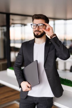 Genç ve modern bir iş adamının portresi dizüstü bilgisayarını tutuyor ve mutlu bir gülümsemeyle kameraya bakıyor.