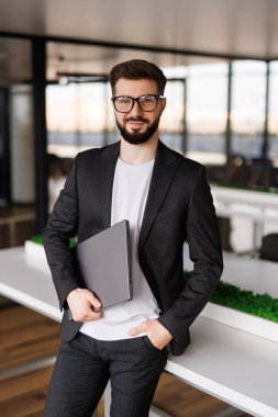 Genç ve modern bir iş adamının portresi dizüstü bilgisayarını tutuyor ve mutlu bir gülümsemeyle kameraya bakıyor.