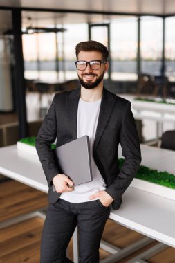 Genç ve modern bir iş adamının portresi dizüstü bilgisayarını tutuyor ve mutlu bir gülümsemeyle kameraya bakıyor.