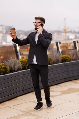 Ofiste kahve fincanı içen gülümseyen işadamı modern işyerinde dururken pencereden bakıyor.