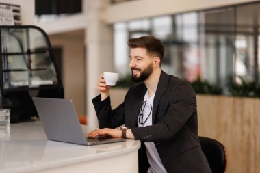 Bir fincan taze kahveden esinlenilmiş. Dizüstü bilgisayarda çalışan ve ofisinde kahve fincanı tutan kendine güvenen genç adam.