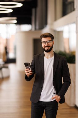 Başarılı yatırımcı ofis içinde çalışır, iş adamı takım elbiseli iş adamı pencere kenarındaki telefonu kullanır, adam gülümser ve akıllı telefondan iyi haberler okur..