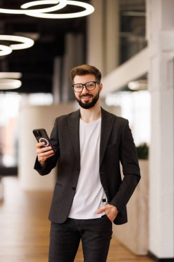 Başarılı yatırımcı ofis içinde çalışır, iş adamı takım elbiseli iş adamı pencere kenarındaki telefonu kullanır, adam gülümser ve akıllı telefondan iyi haberler okur..