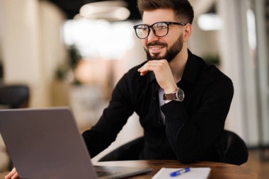 Gözlüklü ve kot gömlekli gülümseyen genç bir adam ofiste oturuyor, internette bir video görüşmesinde çalışıyor ve konuşuyor..
