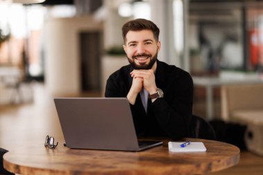 Gözlüklü ve kot gömlekli gülümseyen genç bir adam ofiste oturuyor, internette bir video görüşmesinde çalışıyor ve konuşuyor..