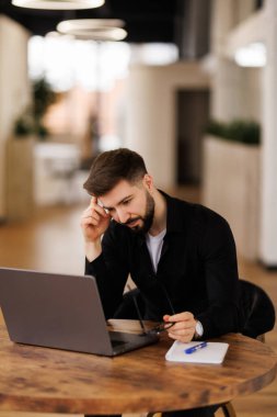 Düşünceli genç yönetici dizüstü bilgisayarda çalışıyor, çalışma masasında oturuyor, ekrana bakıyor, daktilo ediyor, çeneye dokunuyor, çevrimiçi içeriği düşünüyor, iş iletişimi için bilgisayarı kullanıyor.