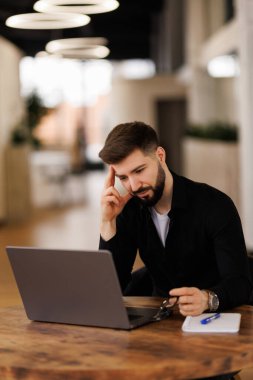 Düşünceli genç yönetici dizüstü bilgisayarda çalışıyor, çalışma masasında oturuyor, ekrana bakıyor, daktilo ediyor, çeneye dokunuyor, çevrimiçi içeriği düşünüyor, iş iletişimi için bilgisayarı kullanıyor.