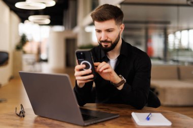 Gülümseyen genç bir ofis çalışanı ofiste oturuyor ve cep telefonu kullanıyor, bir mesaj yazıyor, mektuplaşıyor..