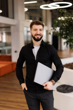 Modern bir ofiste arka planda iş arkadaşlarıyla birlikte dizüstü bilgisayarıyla yürüyen gülümseyen genç bir işadamı.