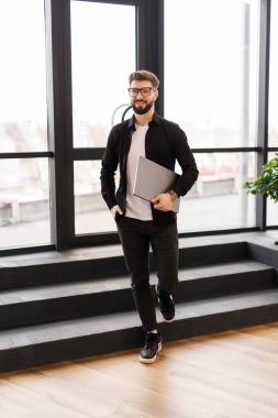 Modern bir ofiste arka planda iş arkadaşlarıyla birlikte dizüstü bilgisayarıyla yürüyen gülümseyen genç bir işadamı.