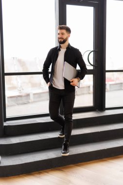 Modern bir ofiste arka planda iş arkadaşlarıyla birlikte dizüstü bilgisayarıyla yürüyen gülümseyen genç bir işadamı.