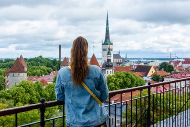 Tallinn, Estonya 'daki Patkuli izleme platformunun arkasındaki kadın, ortaçağ kuleleri, St. Olafs Kilisesi, kırmızı çatılar ve bulutlu gökyüzünün altında Baltık Denizi manzaralı.