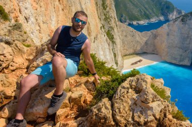 Yunanistan 'ın Zakynthos kentindeki ünlü Navagio Sahili üzerindeki kayalık arazide bir adam dinleniyor. Altında İyon Denizi 'nin ikonik gemi enkazı ve turkuaz suları yatıyor..