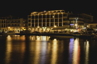 Zakynthos kasabasındaki liman bölgesinin gece görüşü, deniz kenarındaki aydınlık binalar, park edilmiş tekneler ve sakin deniz yüzeyine renkli ışık yansımaları.