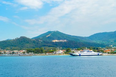 Yunanistan 'ın Thasos adasının limanına bir feribot yanaştı, yeşil bir dağ ve kıyı kasabasının arkaplanı parçalı bulutlu bir yaz gökyüzünün altında..