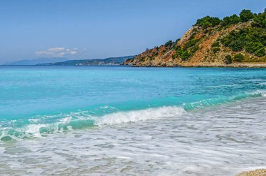 Yunanistan 'ın Kefalonia kentindeki Zola yakınlarındaki Agia Kyriaki plajında turkuaz dalgalar kopuyor. Sakin sular, kızıl kayalıklar, ağaçlar ve huzurlu bir kıyı manzarasında yumuşak bir yaz gökyüzü..