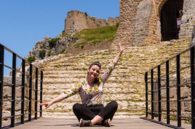 Neşeli bir kadın, Yunanistan 'daki Akrokorint kalesinin taş kapısının önünde kollarını kaldırarak poz veriyor ve tarihi dönüm noktasında güneşli bir günün tadını çıkarıyor..